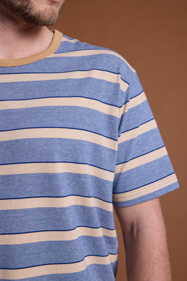 A close-up showing the lower face and upper torso of a person wearing the Barkers Chester Stripe Tee—featuring blue and beige stripes, a beige collar, and soft organic cotton—set against a plain brown background.