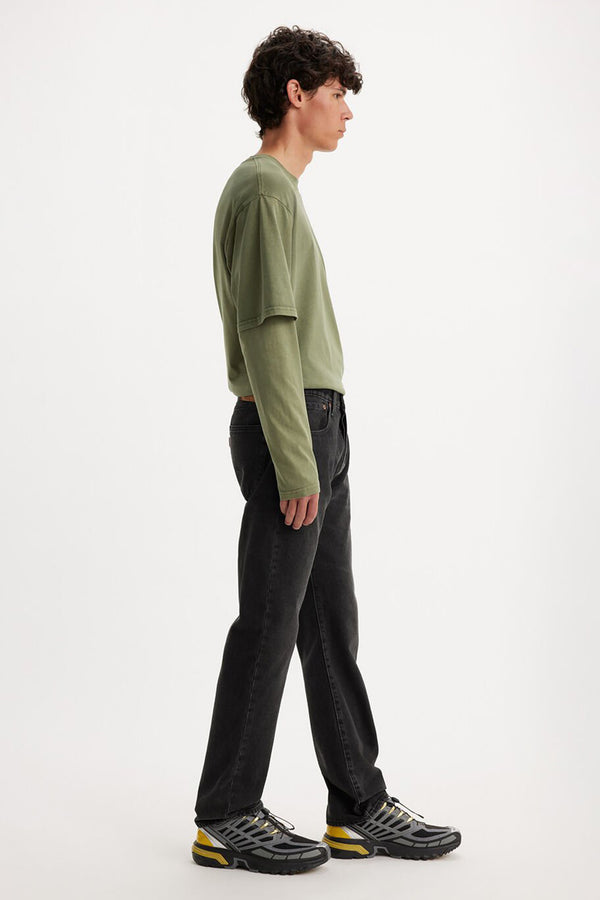 A young man stands in profile against a white background, wearing a loose olive green long-sleeve shirt, Levis 501 Original Jean, and gray running shoes with yellow accents.