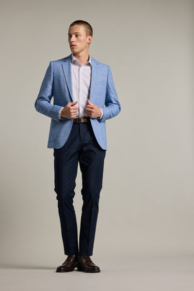A man stands against a plain background wearing the Barkers Mitchel Texture Blazer in light blue, paired with a white dress shirt, navy trousers, and dark brown dress shoes. He looks to the side while adjusting his blazer.