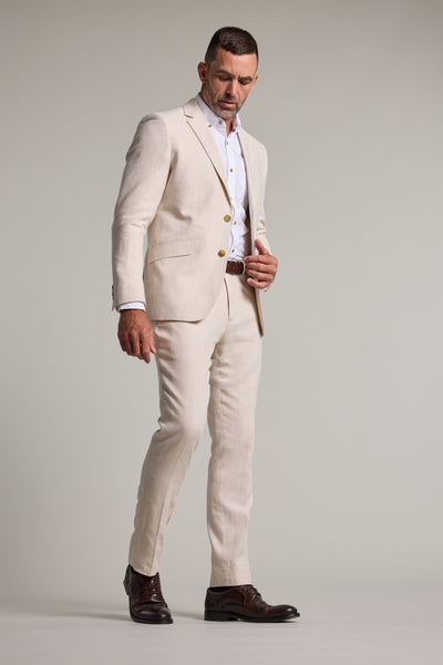 A man stands against a plain background wearing Barkers' Thompson Textured Linen Pant, paired with a white dress shirt, brown belt, and dark brown dress shoes, looking down while adjusting his jacket.