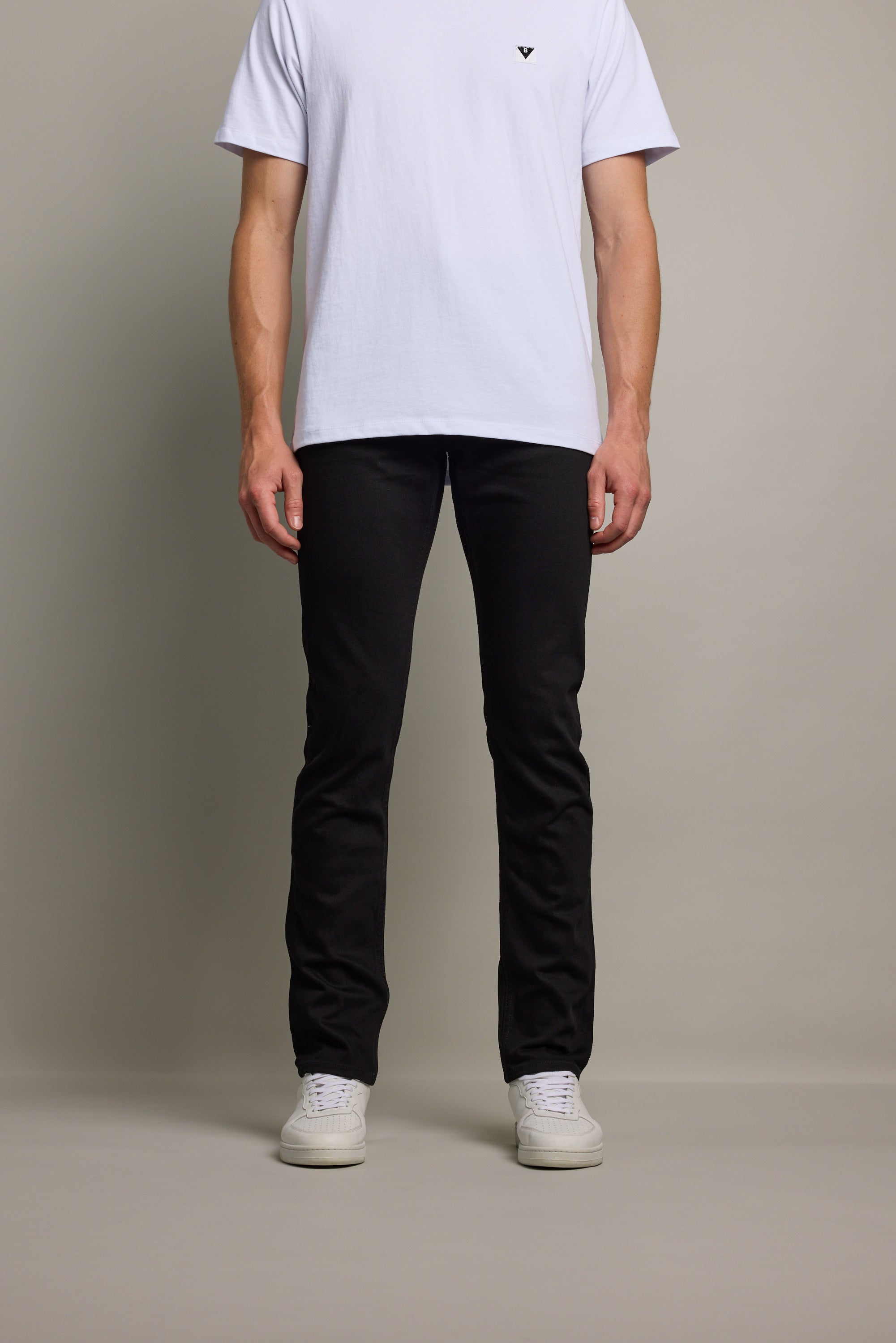 A person, head not visible, stands against a plain background wearing a white t-shirt, Barkers Rambler Denim Slim Jean, and white sneakers.