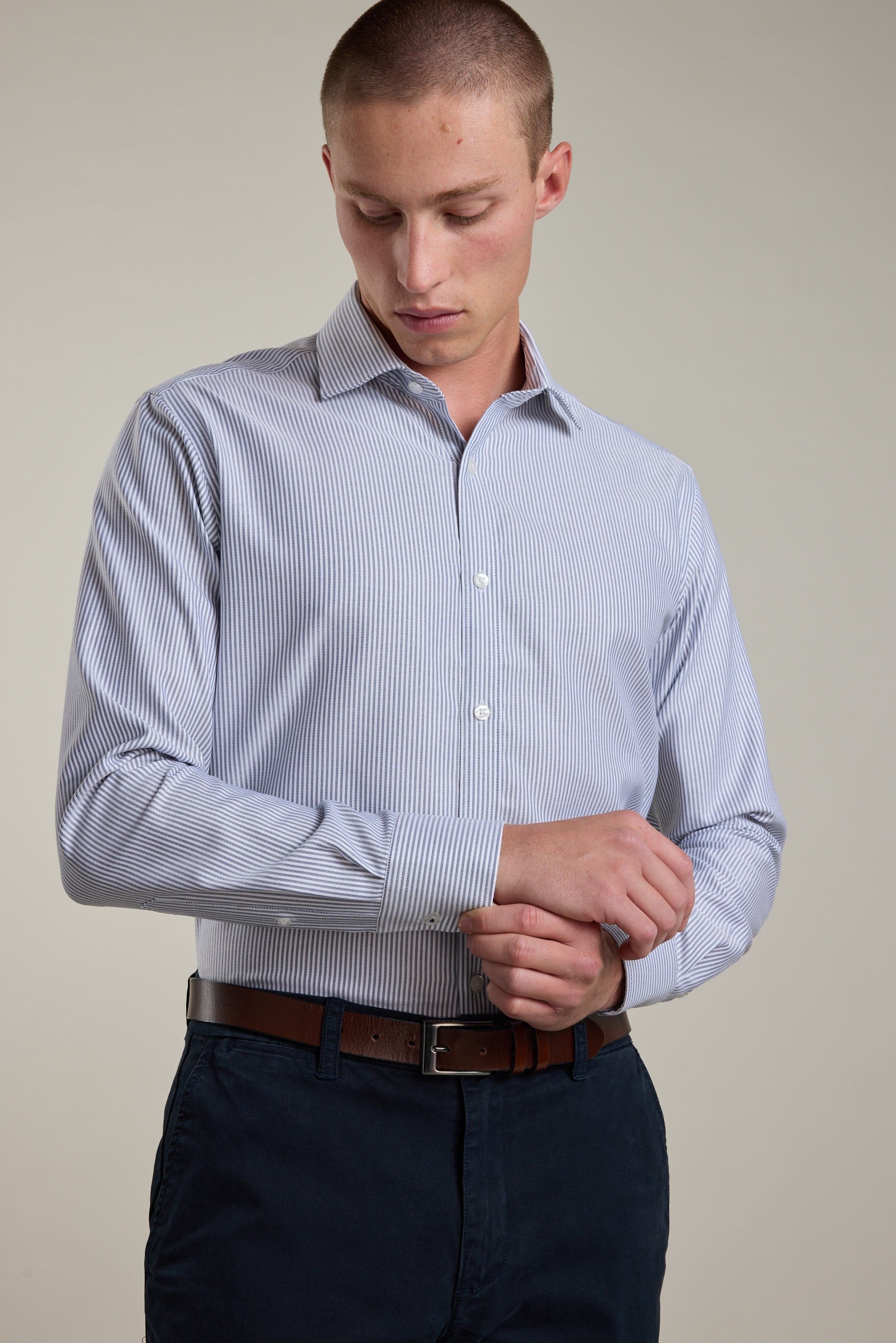 A man stands against a plain background, looking down as he adjusts the cuff of his Barkers Rhea Stripe Business Shirt. The tailored shirt is paired with dark trousers and a brown belt for a polished look.