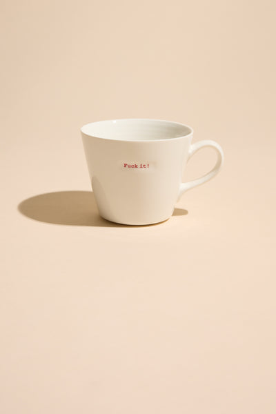 The Bucket Mug - F*ck it! by Other is a white ceramic mug with bold small red text, sitting on a beige surface and casting a shadow—a striking designer piece with attitude.