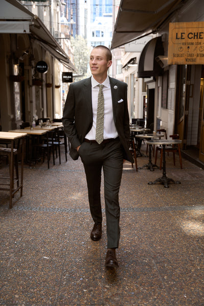 A young man in Barkers' Woodmont Textured Suit Trouser walks confidently down a vibrant city laneway lined with cafés and tables, hands in pockets and a slight smile on his face.
