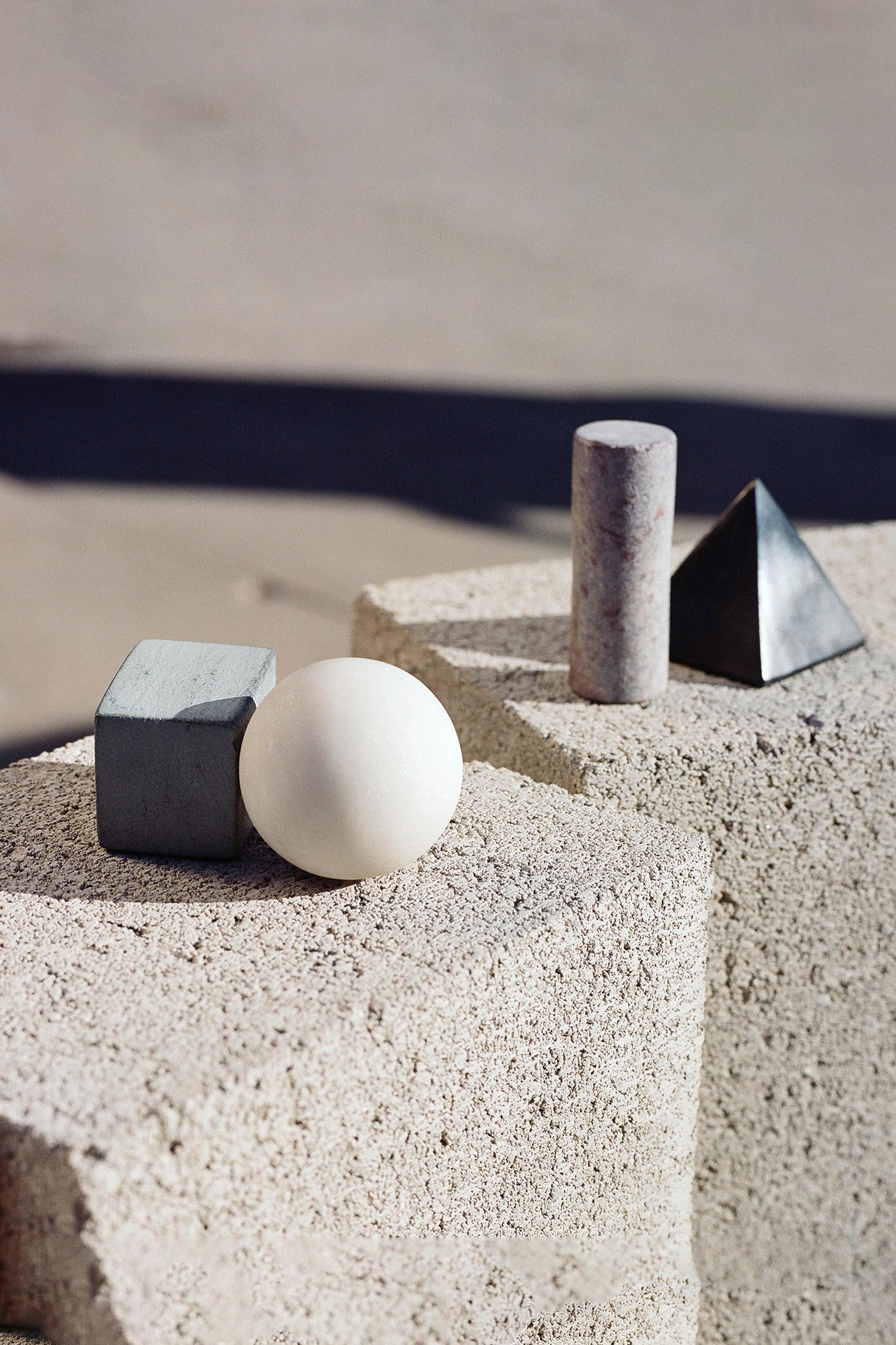 Areaware Drink Rocks by Areaware—a gray cube, white sphere, granite cylinder, and metallic pyramid—rest on light stone surfaces in natural sunlight, evoking elegant cocktail chilling stones.