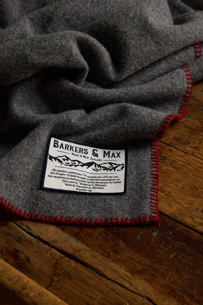 A grey Wool Take Back Blanket by Other, featuring red-stitched edges, lies on a wooden surface. A white label highlights its New Zealand craftsmanship and care details, emphasizing quality and black decorative accents.