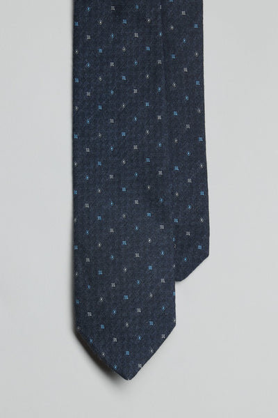 A close-up of the Parisian Ingram Pattern Tie by Parisian, handmade in New Zealand, shows its dark blue geometric print with small blue and white shapes against a plain light background.