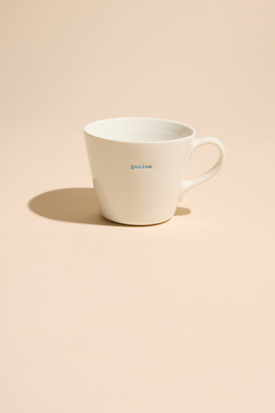 A white ceramic Bucket Mug - Genius by Other, with "genius" in small blue letters, rests on a light beige surface and casts a soft shadow.