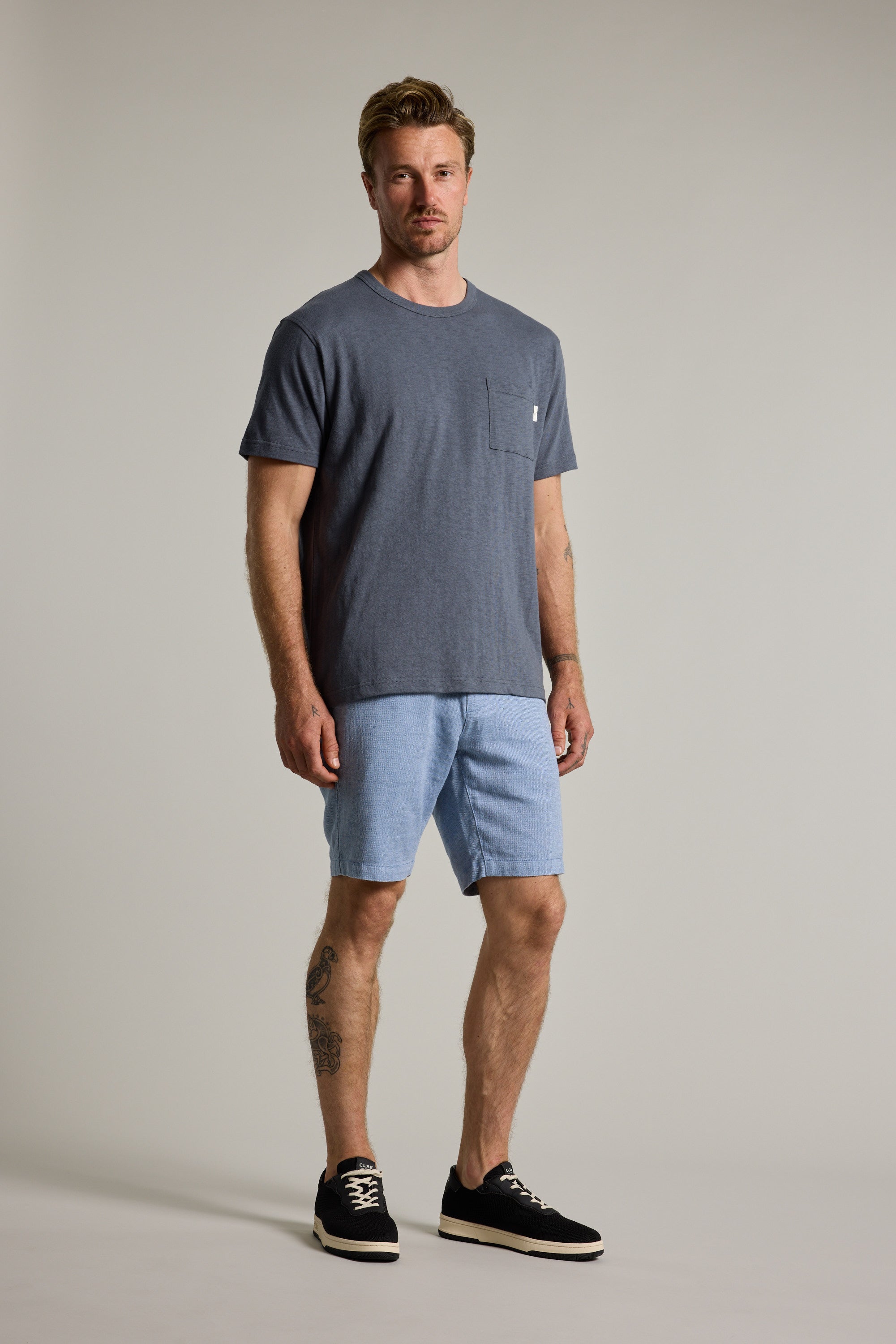 A man faces forward in a dark grey t-shirt, Barkers Elliot Texture Walk Short in light blue, and black sneakers. He has light brown hair, a tattoo on his left calf, and stands against a plain, light background.