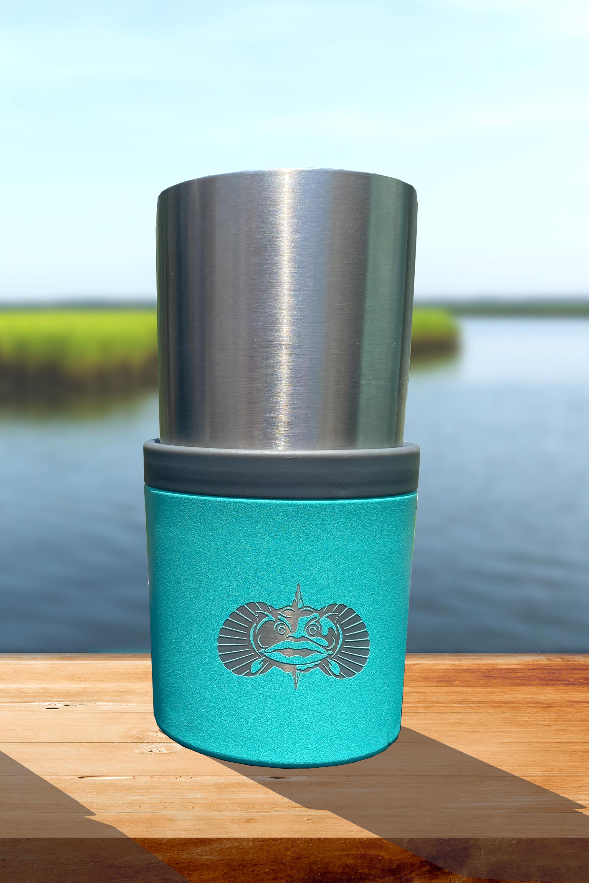 A Toadfish The Anchor non-tipping cup holder in turquoise, featuring a fish logo, holds a silver tumbler and rests on wood outdoors by calm water with green grass behind.