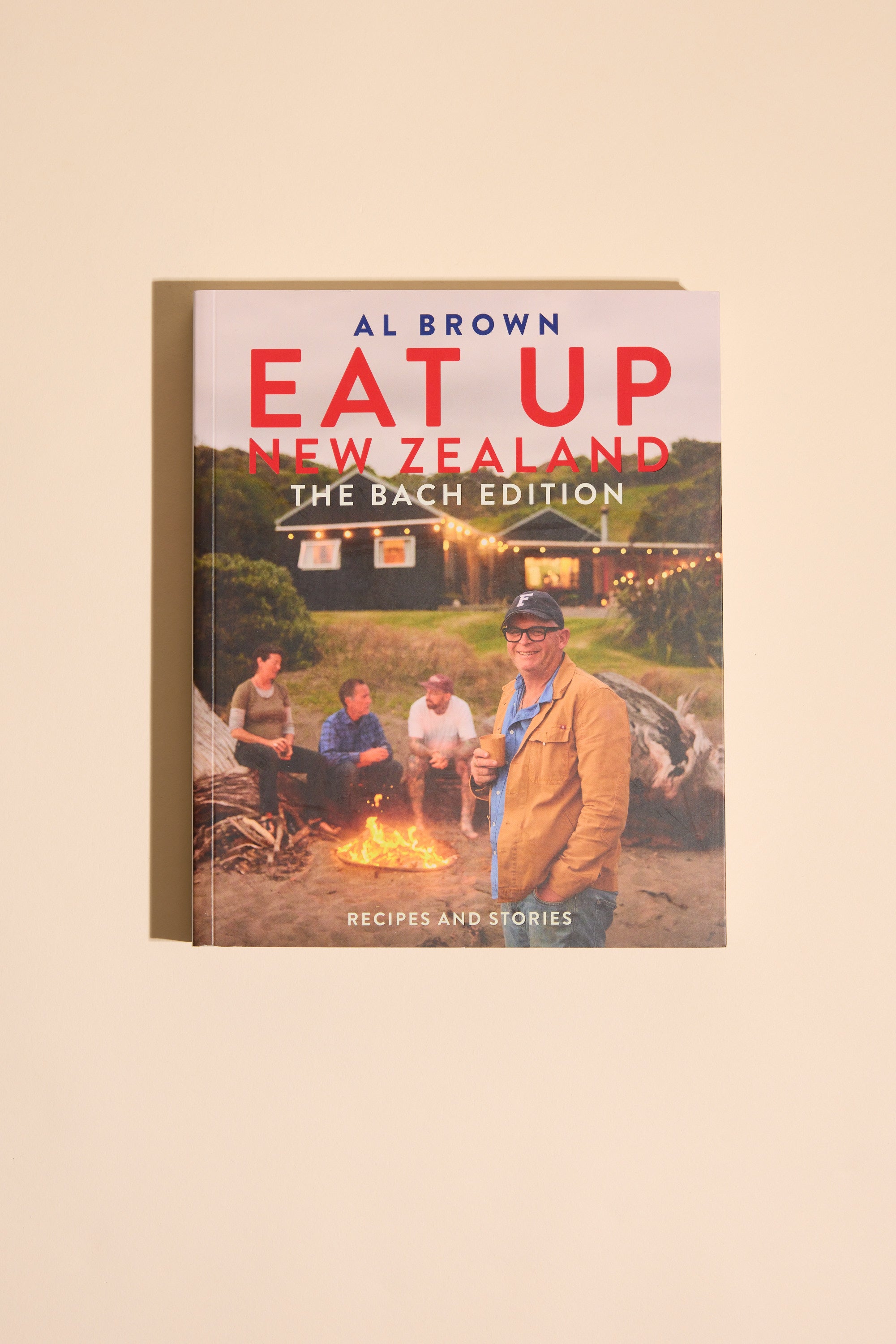 The Other brand’s Eat Up New Zealand The Bach Edition cookbook sits on a beige surface, its cover showing people cooking by a campfire outdoors with a house in the background.