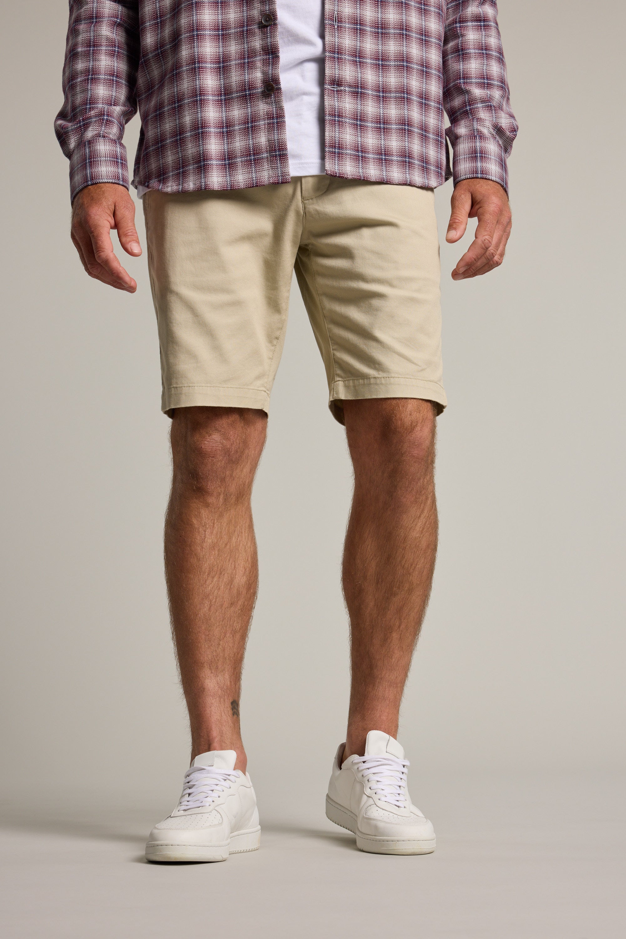 A person, visible only from the waist down, models Barkers' Braddock Walk Short in beige with a purple plaid shirt and white sneakers against a plain background.