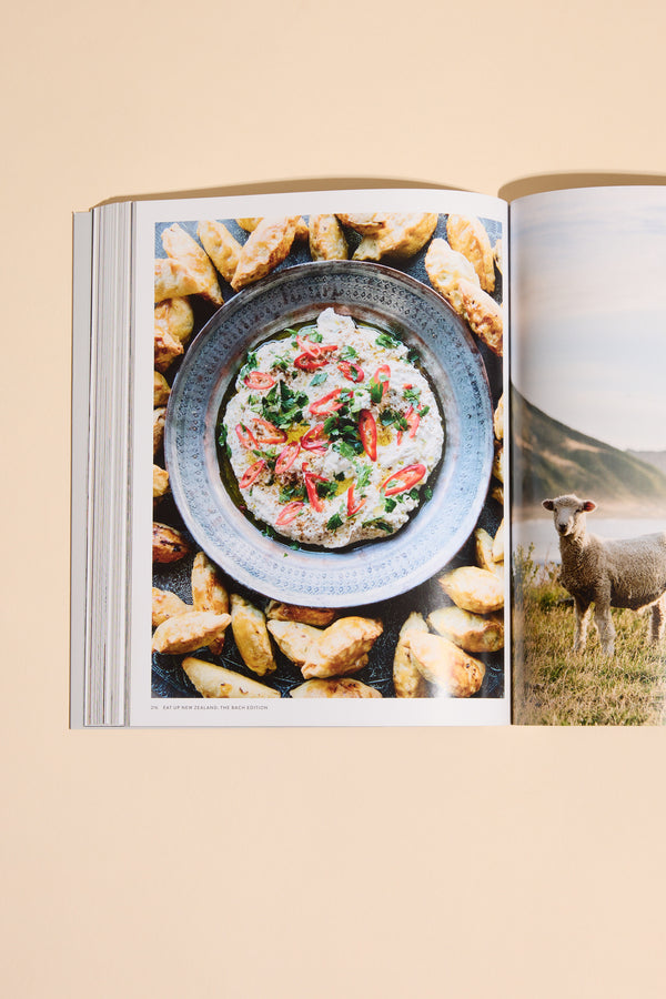 The Eat Up New Zealand The Bach Edition by Other features a cookbook open to creamy dip with herbs and red peppers, surrounded by golden pastries—ideal for bach cooking with seasonal ingredients. Another page shows a sheep in a grassy field.