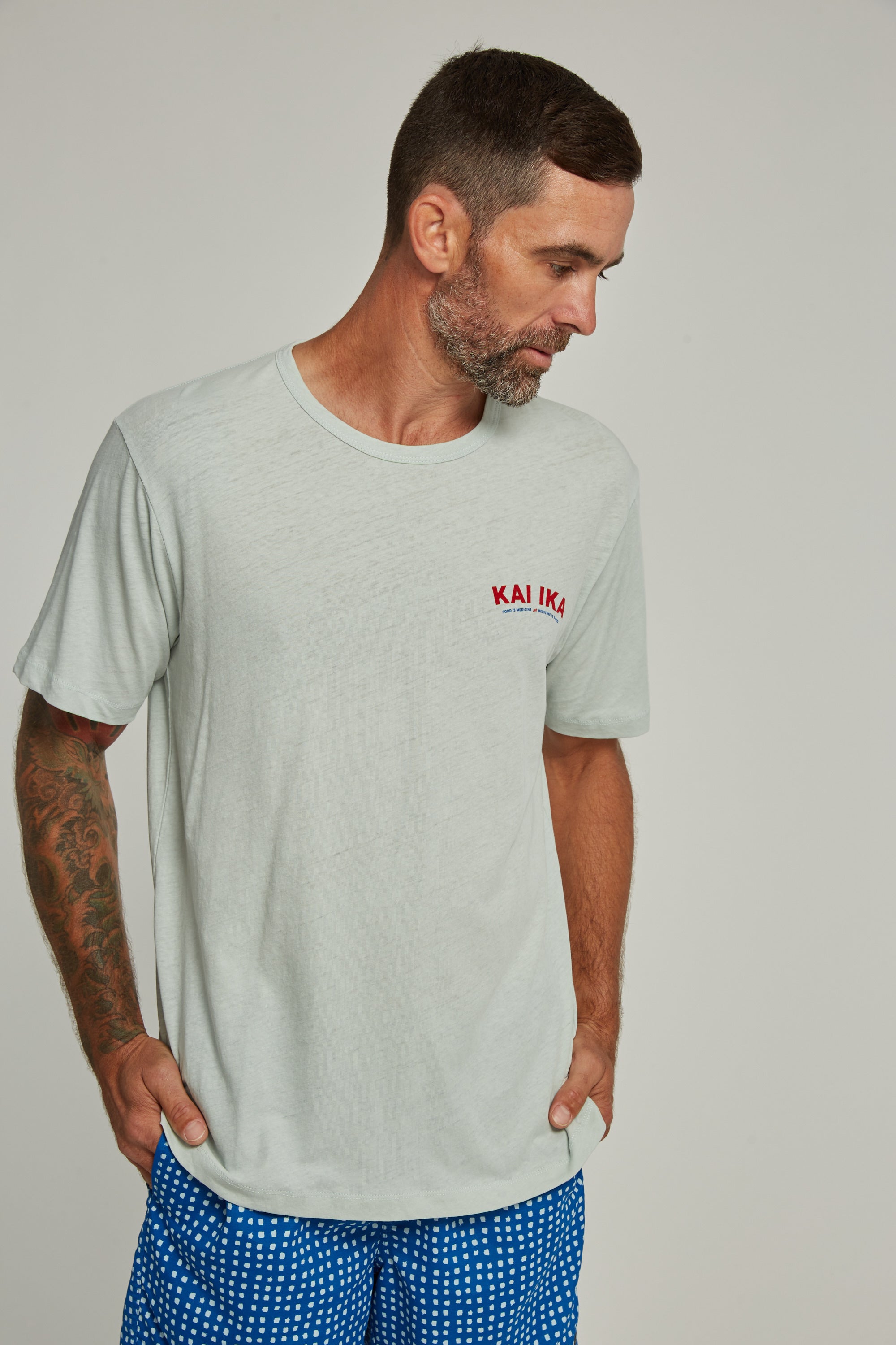 A bearded man with short hair, wearing the Legasea Kai Ika Snapper Tee by Legasea, blue patterned shorts, and showing a right arm tattoo, looks down with his hands in his pockets.