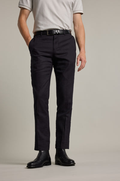 A person stands with hands in pockets against a plain light background, wearing Barkers Windsor Texture Linen Pant in a slim tapered fit, paired with a light gray short-sleeve polo shirt, black belt, and black leather boots. Face not visible.