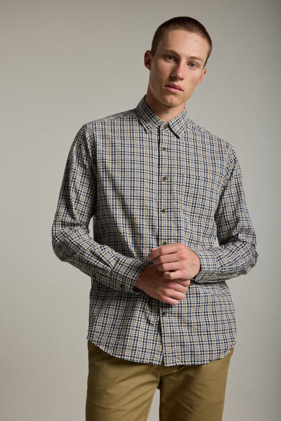 A young man with short hair wears the Barkers Goode Fine Check Shirt in beige and blue cotton, paired with khaki pants, standing against a plain light background with his hands clasped in front.