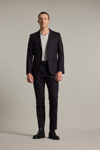 A man stands against a plain background, wearing Barkers' Windsor Texture Linen Blazer over a light shirt, paired with a black belt and black dress shoes. He faces forward with a neutral expression and relaxed posture.