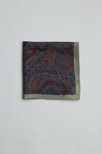 The Parisian Paisley Wool PS by Parisian is a folded wool pocket square with paisley print in red, blue, and purple tones, bordered by muted green, set on a plain light background. Expertly crafted in New Zealand.