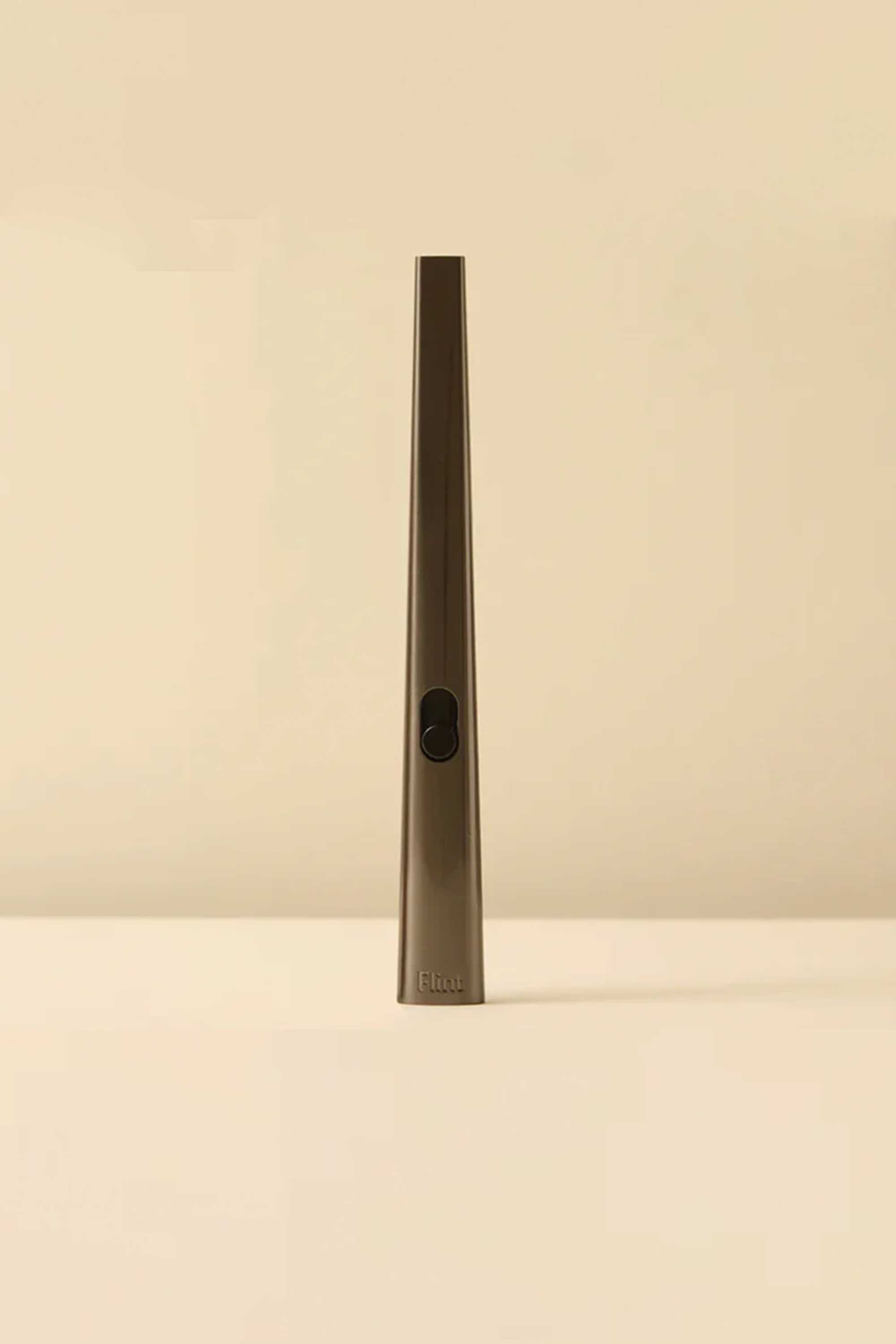 The Flint Rechargeable Lighter 2.0 by Flint is a slim, metallic, wand-shaped gold lighter with a black button, standing upright on a flat beige surface against a matching beige background.