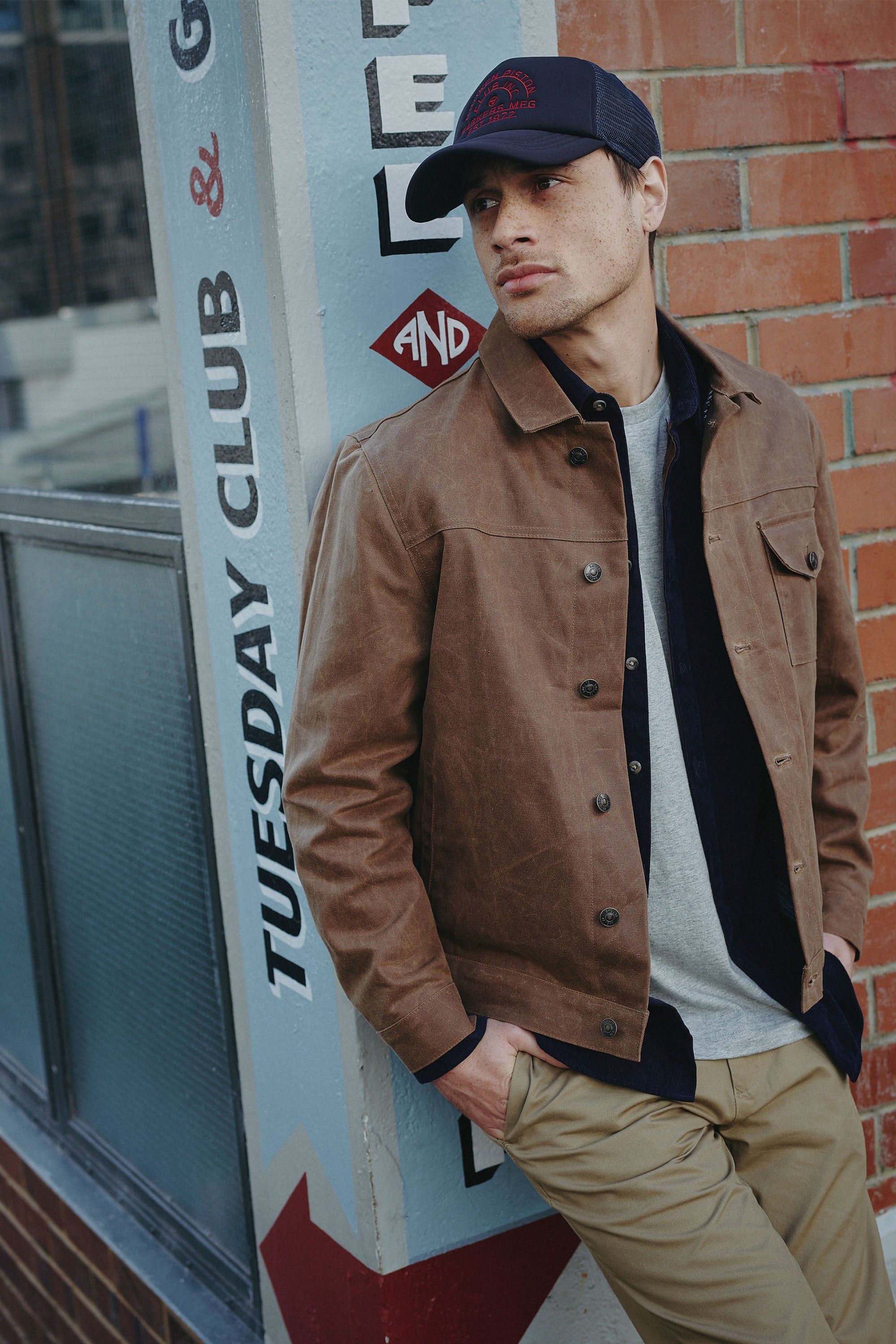 Wearing the Barkers Piston Club Trucker Cap with an embroidered logo, a man leans against a sign and highlights its adjustable fit.