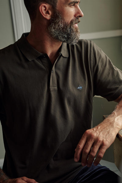 A bearded man sits indoors with one arm on a wooden surface, wearing the Barkers Regent Pique Polo in dark olive, and looks off to the side.