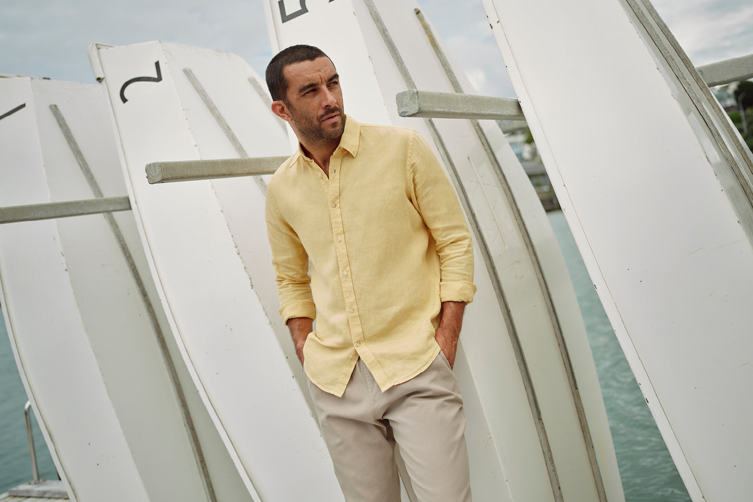 A man in a yellow button-down shirt and light pants stands with hands in pockets beside upright white rowboats by the water, looking thoughtfully into the distance.