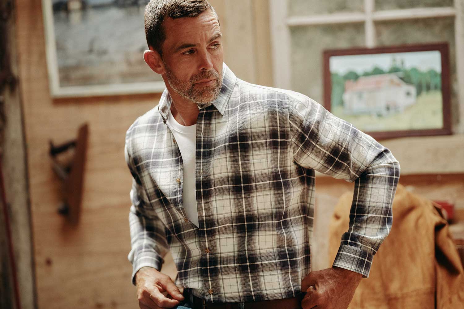 A man in a yellow button-up shirt and beige pants stands in front of white boats lined up vertically by the water, looking to the side with his hands in his pockets.