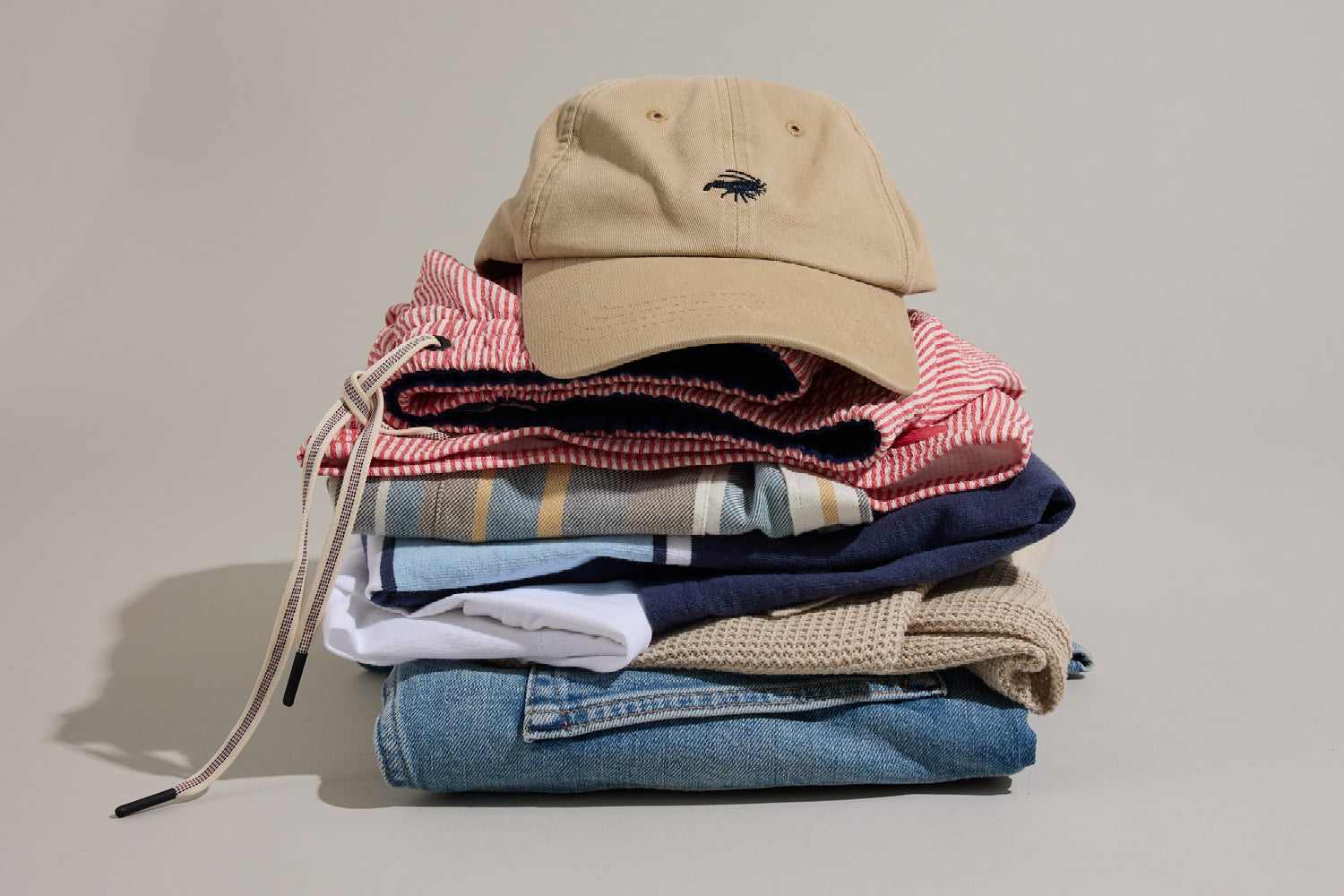 A neatly folded stack of casual clothes including jeans, shirts, and a red striped garment, topped with a beige baseball cap with a small embroidered logo, all set against a neutral background.