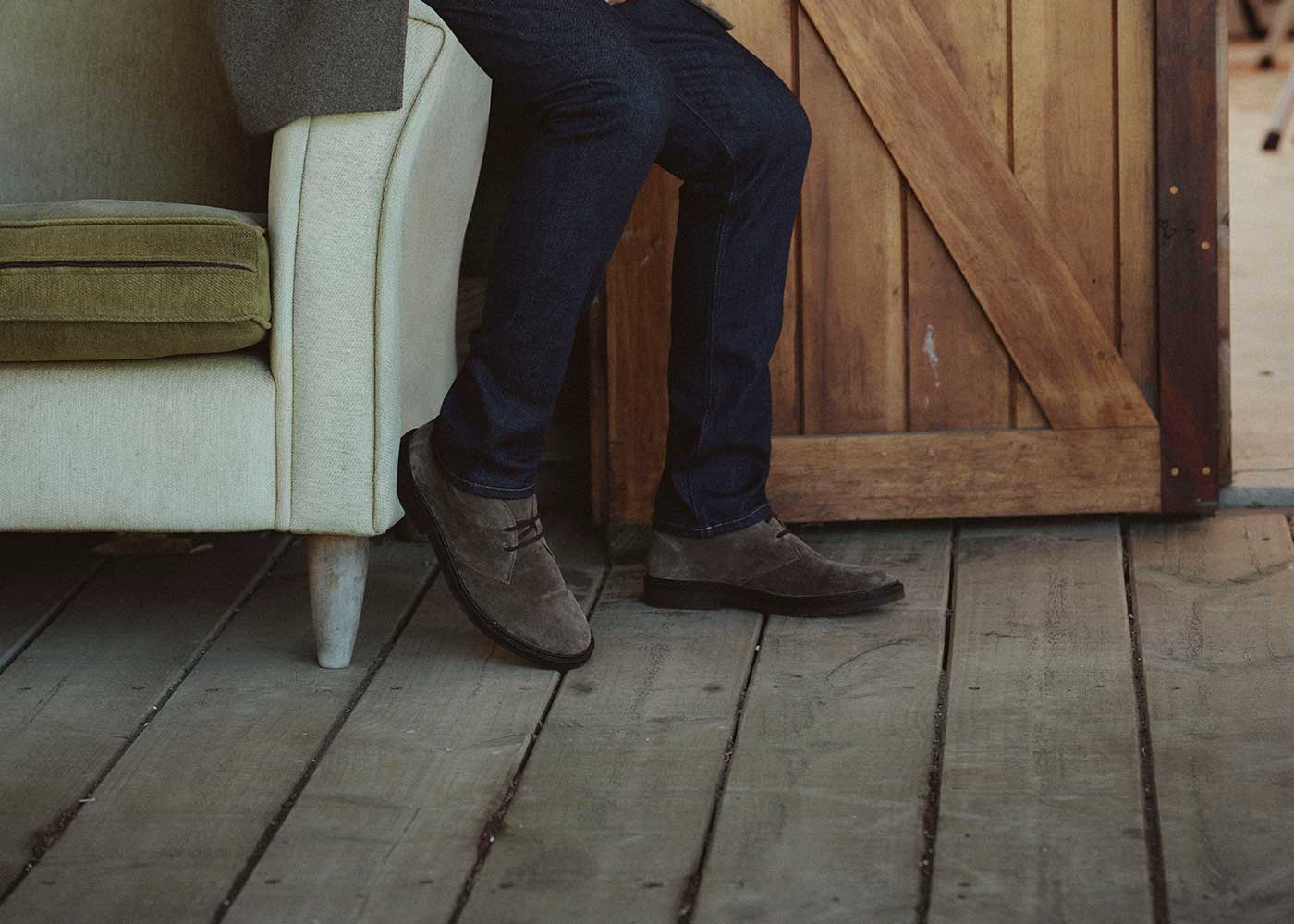 A person wearing dark jeans and brown suede shoes sits on a light green sofa next to a wooden door on a rustic wooden floor. Only the lower half of their body is visible.