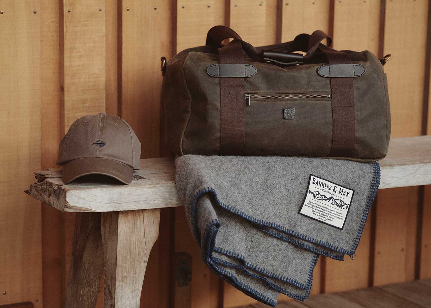 A brown duffel bag, a taupe baseball cap, and a folded gray wool blanket with a label sit on a rustic wooden bench against a wood-paneled wall.