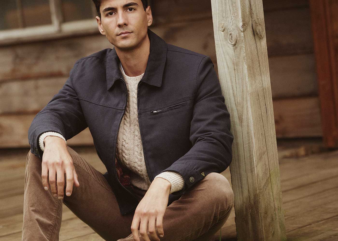 A man in a navy jacket and beige sweater sits on a wooden porch, leaning against a post, with a rustic wooden building in the background.