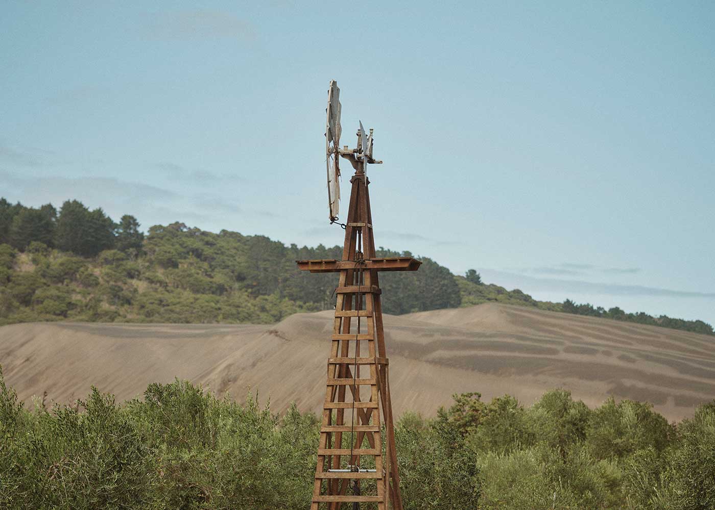 A rustic wooden windmill stands among green shrubs, with rolling hills and a forested area in the background under a partly cloudy sky.