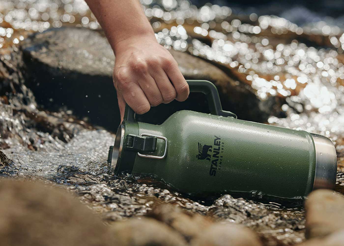 A hand holds a green Stanley thermos jug, partially submerged in a flowing stream with rocks visible in the water.