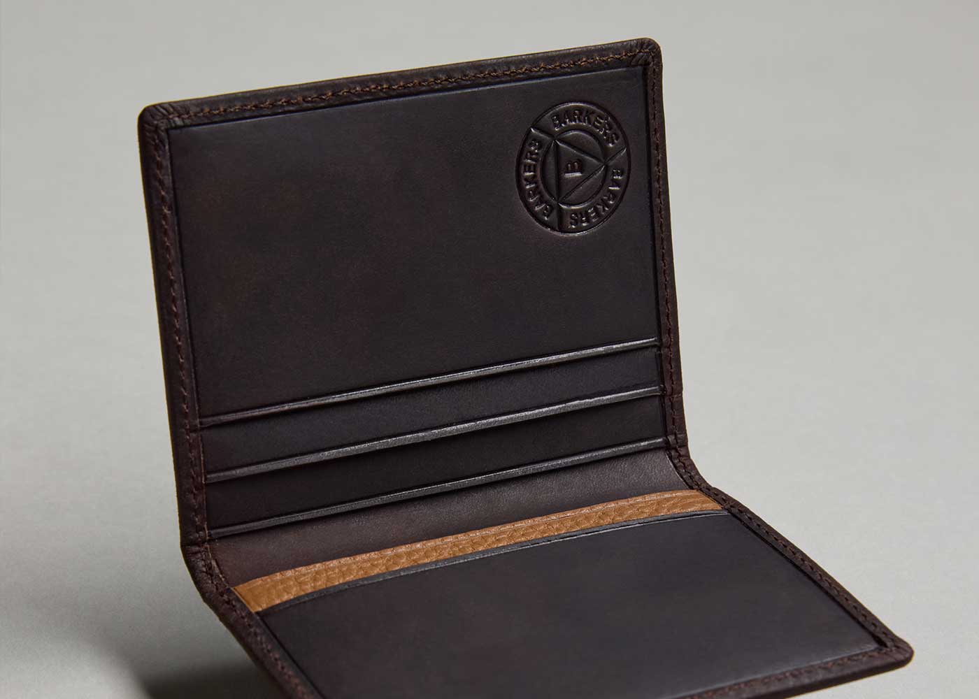 A close-up of an open dark brown leather wallet with card slots and a circular embossed logo on the inside. The wallet is empty and placed on a plain gray background.