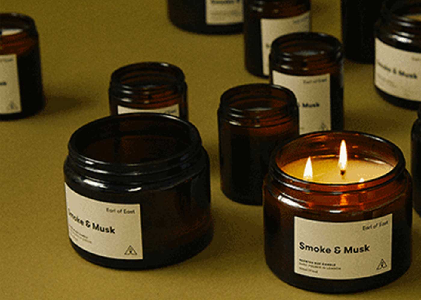 Several brown glass jars labeled Smoke & Musk are displayed on a surface. One jar contains a lit candle with two flames, while the others have unlit candles or lids.