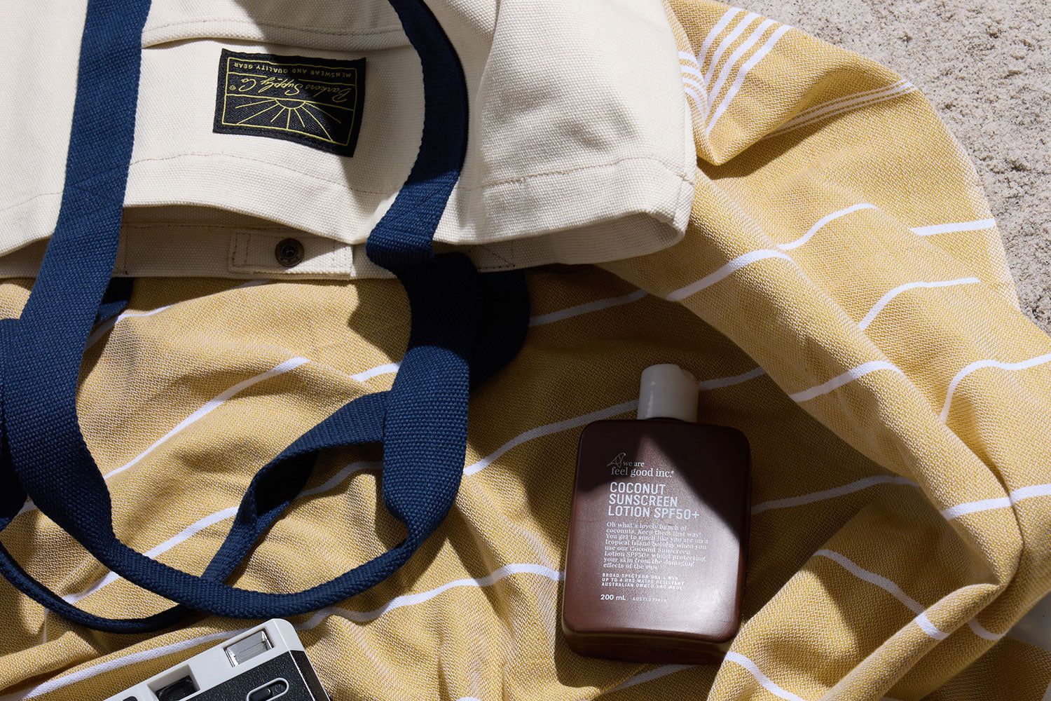A bottle of coconut sunscreen lotion lies on a yellow striped towel next to a beige tote bag with navy blue straps, set on a sandy surface.