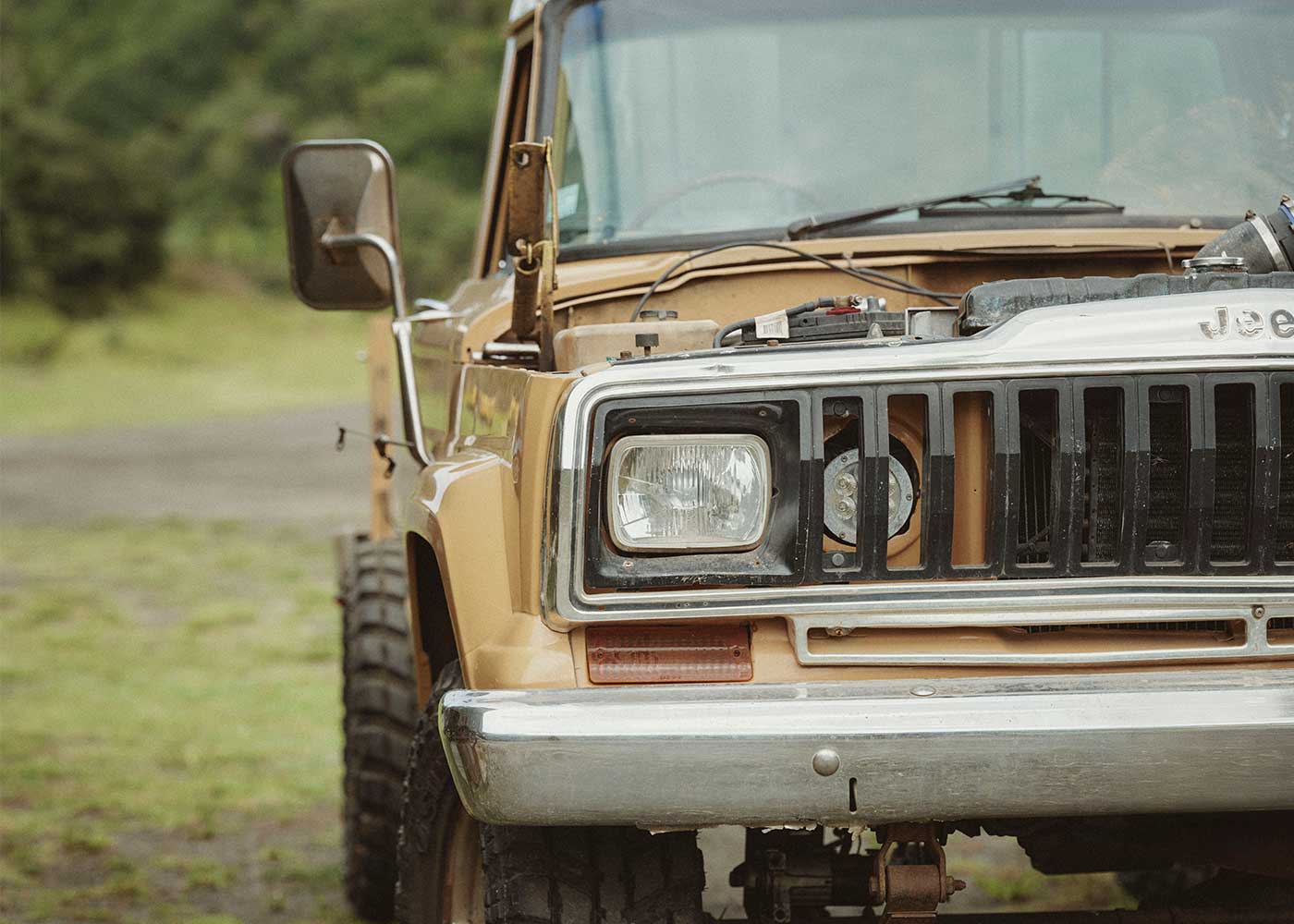 Close-up of the front of a vintage tan Jeep truck with its hood open, showing parts of the engine. The vehicle is parked outdoors on grass, with greenery in the background.