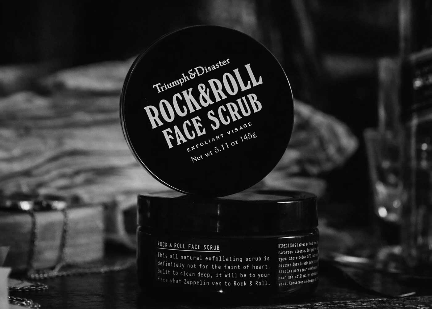A black jar of Triumph & Disaster Rock & Roll Face Scrub with the lid leaning against it, featuring bold white text. The background is blurred with indistinct objects.