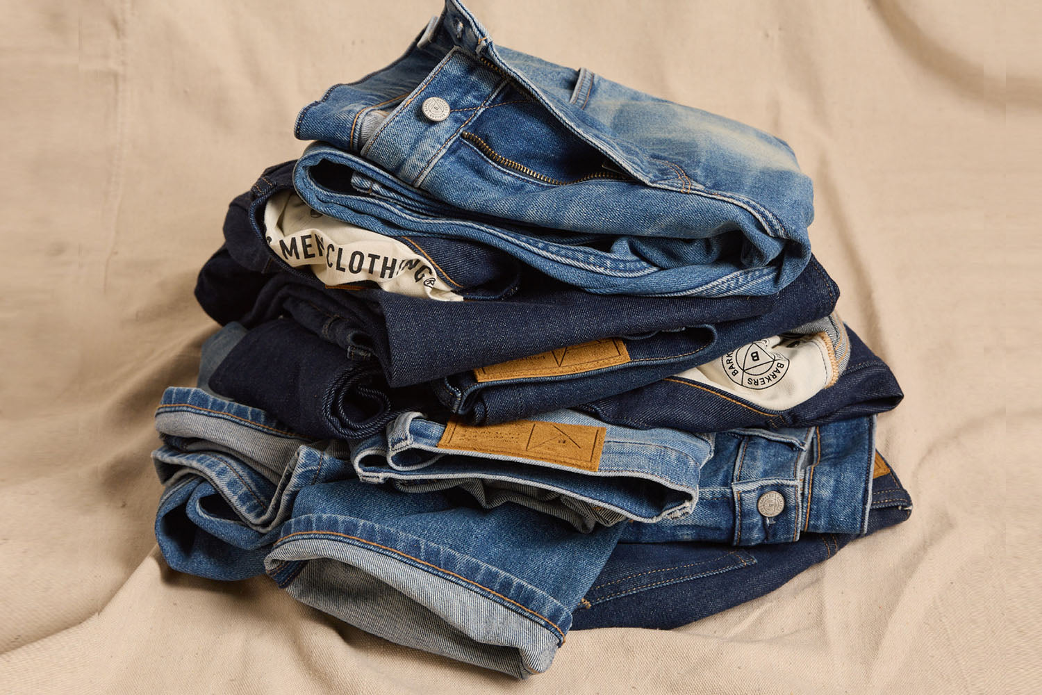 A stack of folded blue jeans and denim jackets in various shades of blue lying on a beige fabric surface. Some have visible leather patches and branded labels.