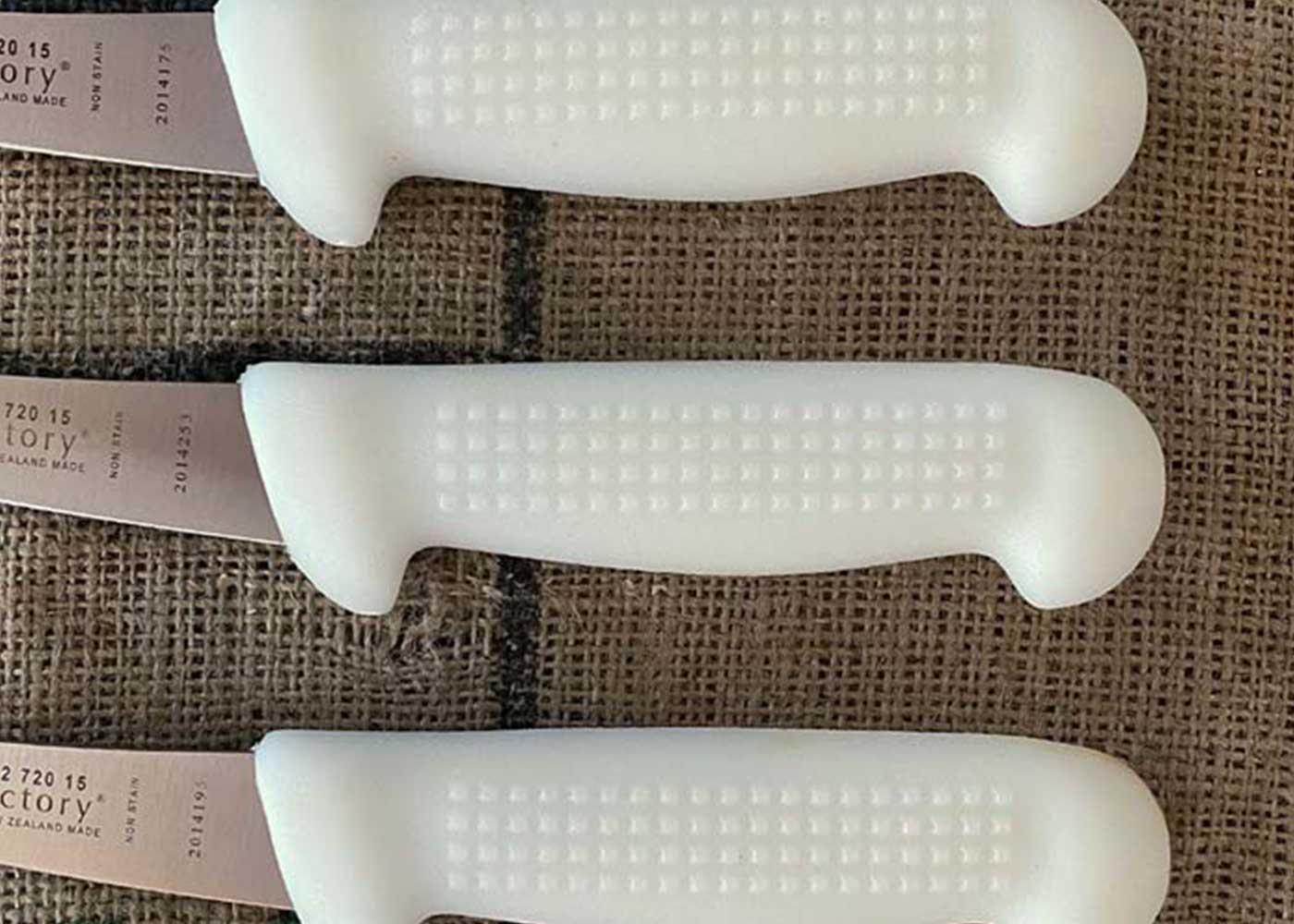 Three kitchen knives with white textured handles are arranged parallel to each other on top of a burlap surface.
