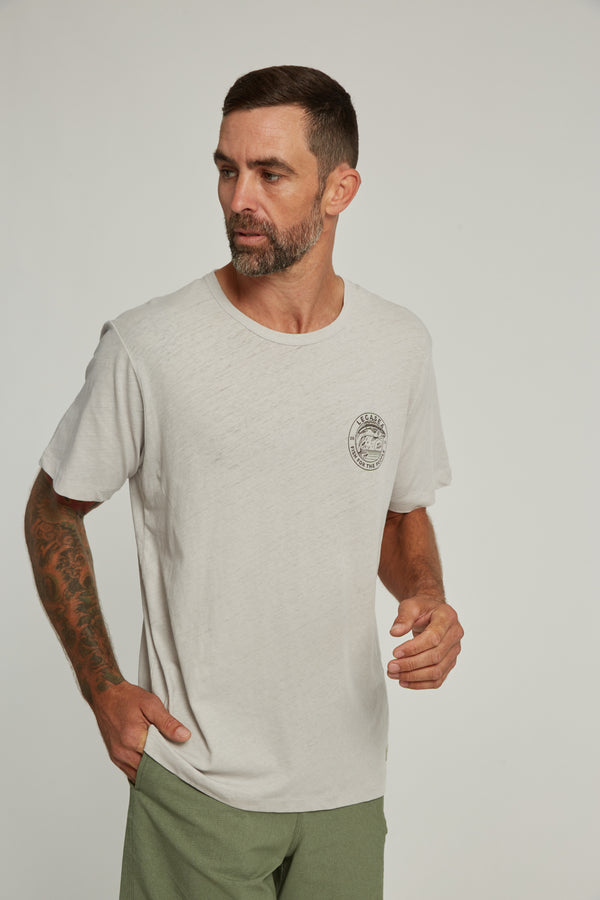 A man with short dark hair and a beard wears the Legasea Stamp Tee by Legasea and green pants, standing against a plain background, looking right with one hand raised.