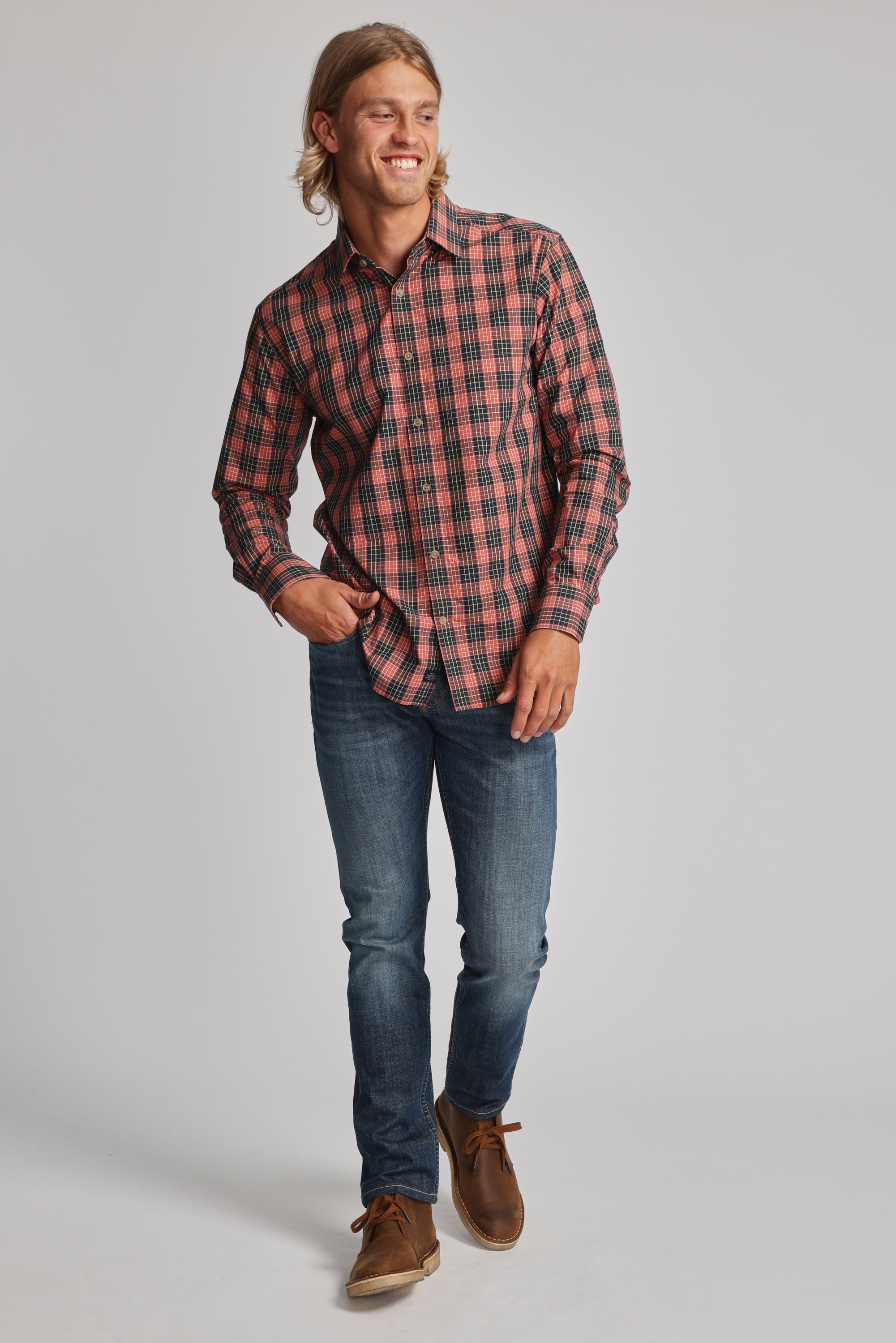 A smiling man with long blond hair wears the Barkers Formby Long Sleeve Shirt in red and black plaid, paired with blue jeans and brown shoes. He stands against a light gray background, one hand in his pocket and the other holding his shirt.