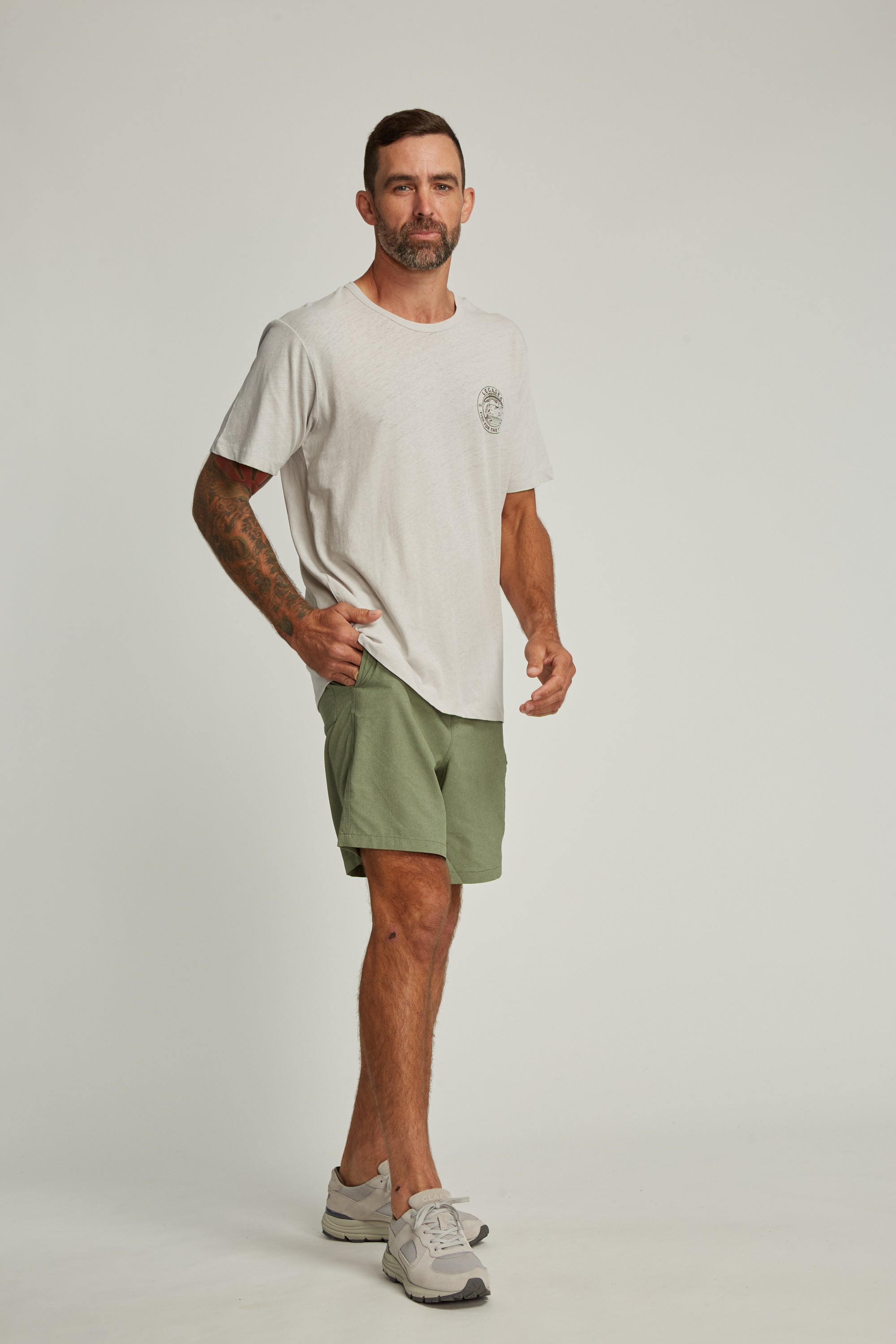 A man with short hair, a beard, and arm tattoos poses against a plain background wearing the Legasea Stamp Tee by Legasea, styled with green shorts and grey sneakers. One hand rests on his hip.