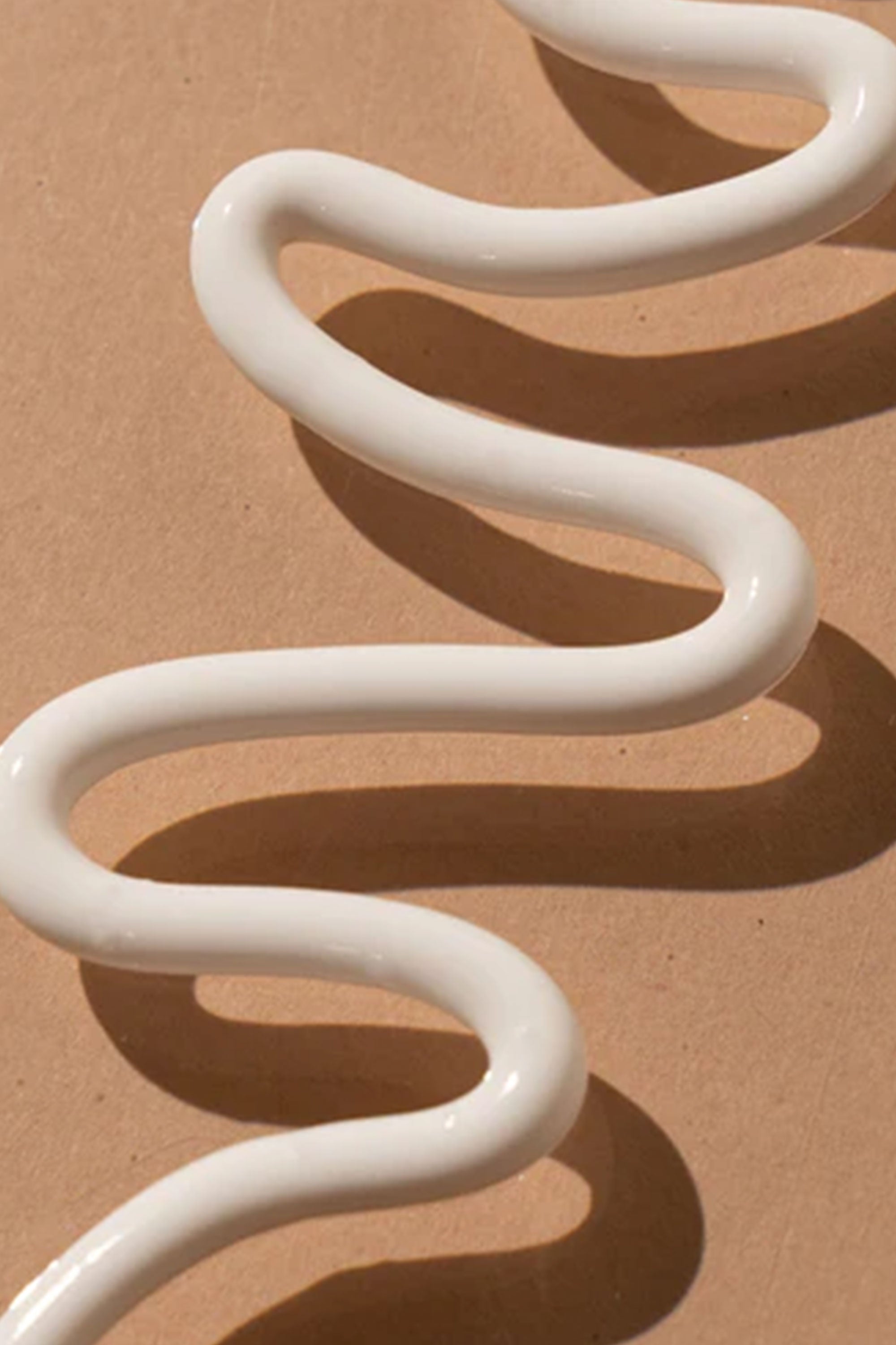A thick, glossy white line of Other Coconut Sunscreen Lotion SPF50+ 75ml (Traveller) is arranged in a wavy, zigzag pattern on a light brown surface, highlighting its broad spectrum protection.