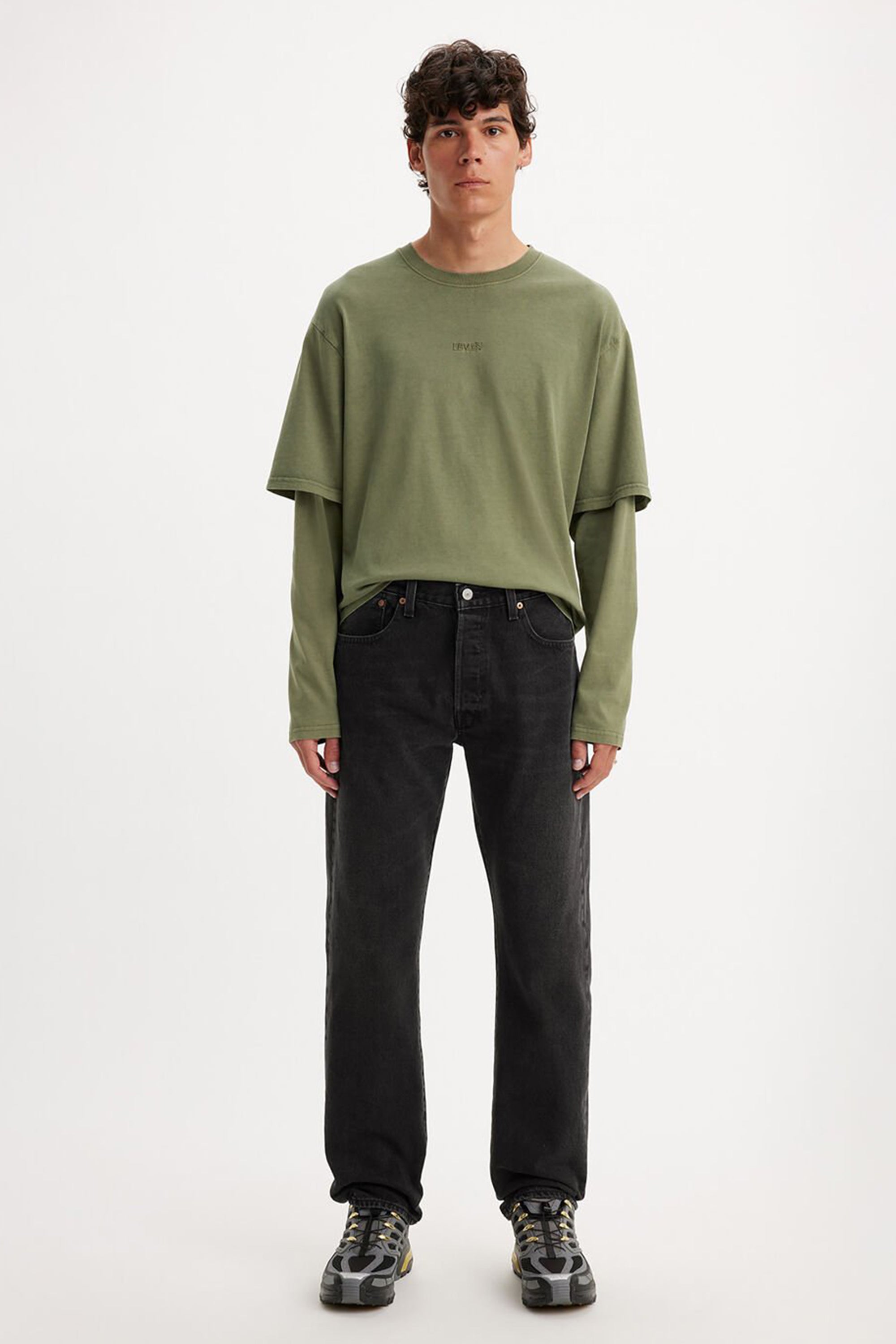 A person faces forward against a plain white background, wearing layered green shirts, Levis 501 Original Jean straight-leg jeans, and black-and-gray sneakers.