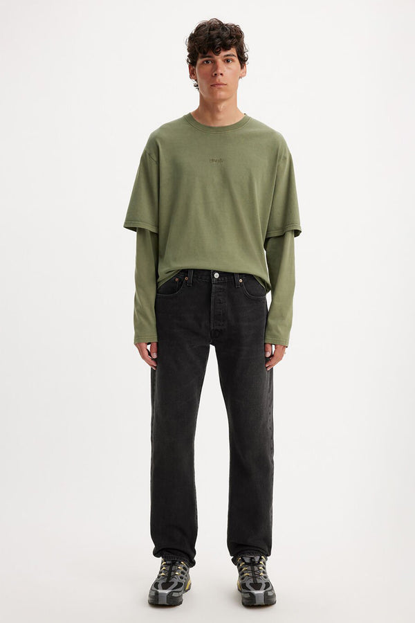 A person faces forward against a plain white background, wearing layered green shirts, Levis 501 Original Jean straight-leg jeans, and black-and-gray sneakers.