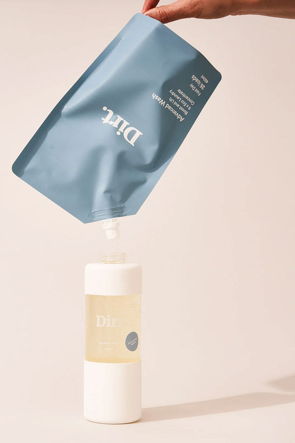 A hand pours laundry detergent from a blue Other Advanced Wash Starter Pack pouch into a clear, white-based bottle with matching branding, against a neutral background—ideal for odour removal.