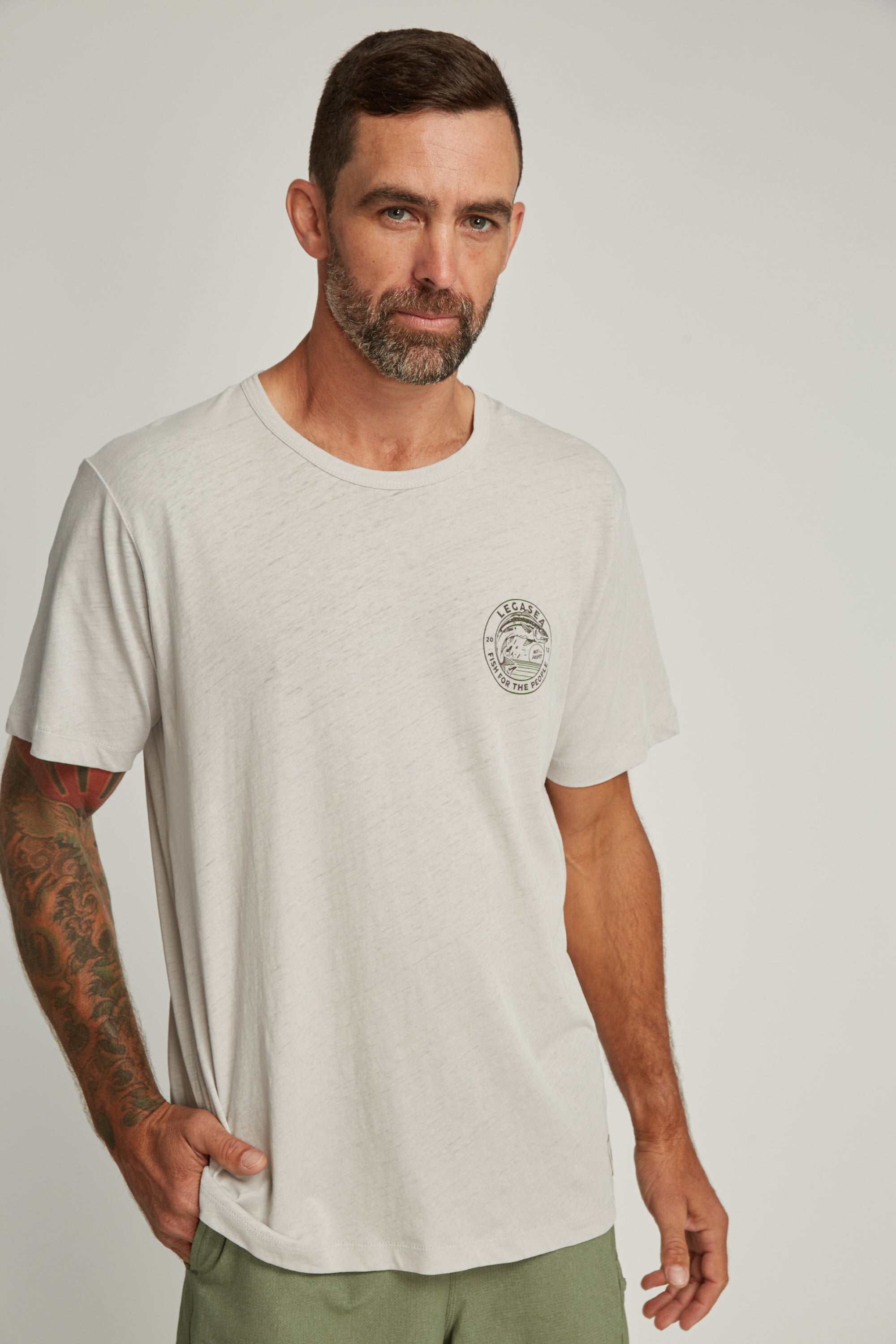 A man with short dark hair, a beard, and a tattooed right arm wears the Legasea Stamp Tee by Legasea, paired with green pants, standing against a plain light background.