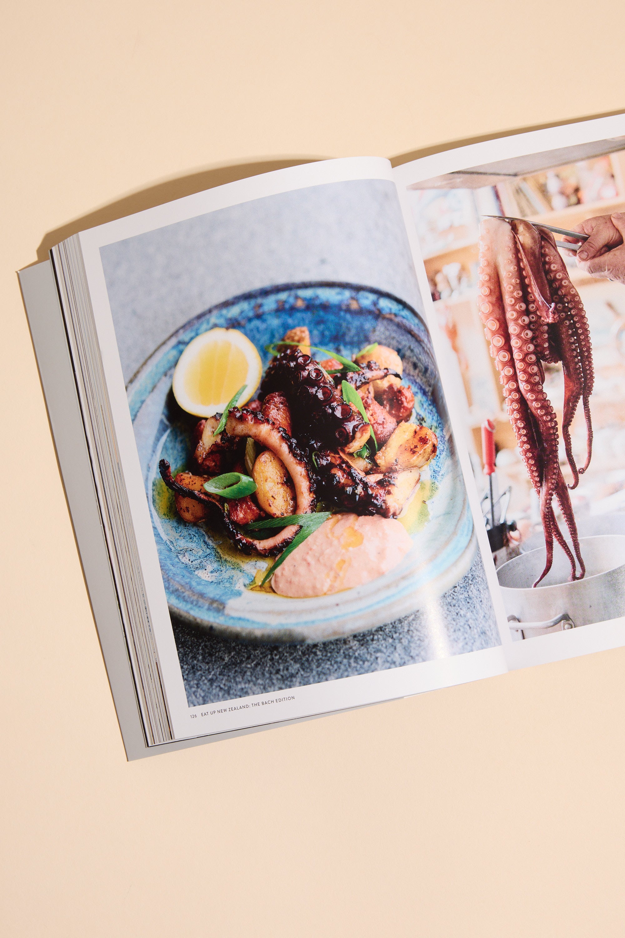 An open Eat Up New Zealand The Bach Edition cookbook by Other features grilled octopus with lemon and sauce on one page, and a hand holding raw octopus on the other—ideal for bach cooking or trying New Zealand recipes.