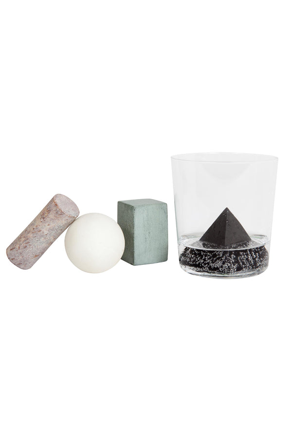 Areaware Drink Rocks by Areaware—featuring a gray cylinder, white sphere, and greenish cube—are lined up beside a clear glass holding a black pyramid on dark granules. All items rest on a white background.